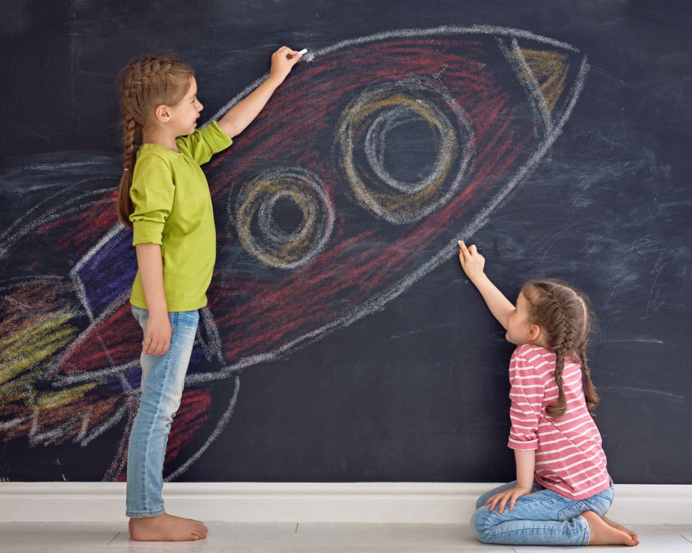 Dos niñas dibujando en una pared de pizarra un cohete.