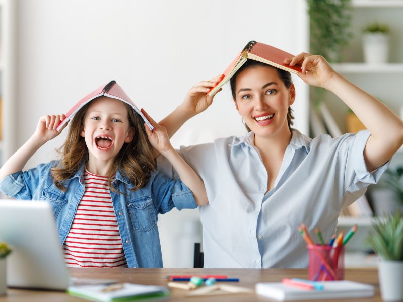 Madre e hija sentadas haciendo los deberes con el cuaderno encima de la cabeza. Imagen que representa técnicas de estudio emocionante.